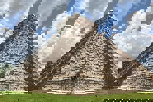 Temple of Kukulcan pyramid at Chichen Itza, Yucatan, Mexico, with cloudy blue sky and green grass foreground. 