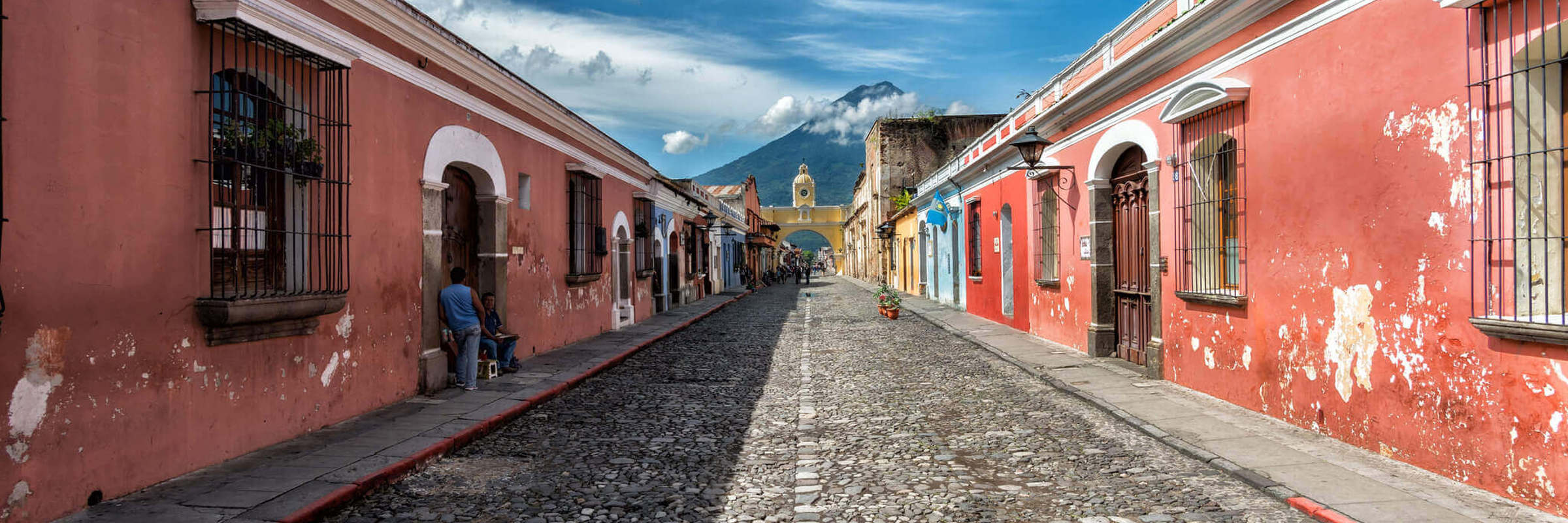 Cobblestone street in Antigua, Guatemala with pastel houses and a distant volcano under blue skies. 
